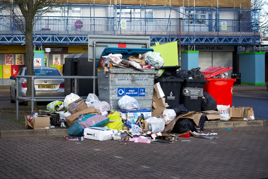 A close-up view of a large pile of mixed domestic waste scattered on a pavement or driveway, including crumpled paper, plastic bottles, metal cans, and used tissue or paper towels. The plastic bottles
